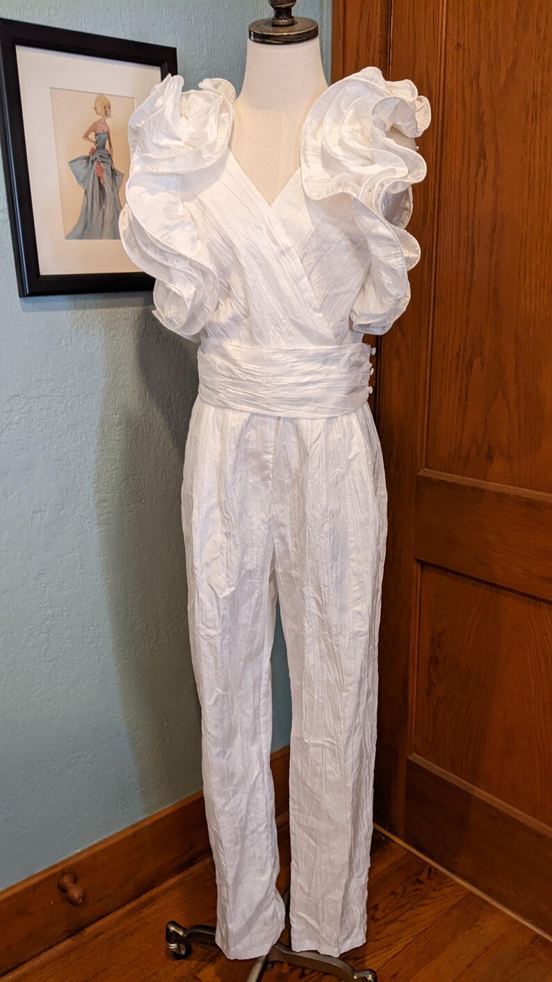 May include: A white jumpsuit with a V-neckline and ruffled sleeves. The waist is cinched with a matching fabric belt. The jumpsuit is displayed on a mannequin. A framed picture hangs in the background.