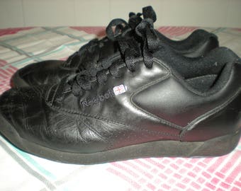 Pode incluir: Um par de ténis Reebok pretos com parte superior em pele lisa, atacadores pretos e um pequeno emblema da bandeira Union Jack na lateral. Os sapatos têm uma silhueta de cano baixo com uma biqueira texturizada e uma sola de borracha preta.