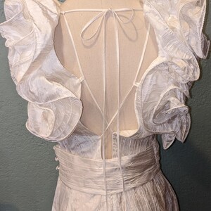 May include: White jumpsuit with ruffled sleeves and a low back. The fabric has a textured appearance, and the waist is cinched with a matching sash. The back features delicate tie details. The garment is displayed on a mannequin.