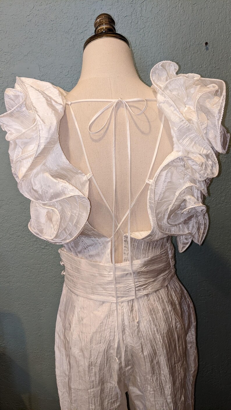 May include: White jumpsuit with ruffled sleeves and a low back. The fabric has a textured appearance, and the waist is cinched with a matching sash. The back features delicate tie details. The garment is displayed on a mannequin.
