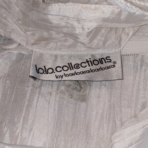 May include: Close-up of a white garment with a textured, pleated fabric. The label reads "bb.collections by barbarabarbarara" with a size 12 tag. The fabric appears to be lightweight and flowing, with a subtle sheen.