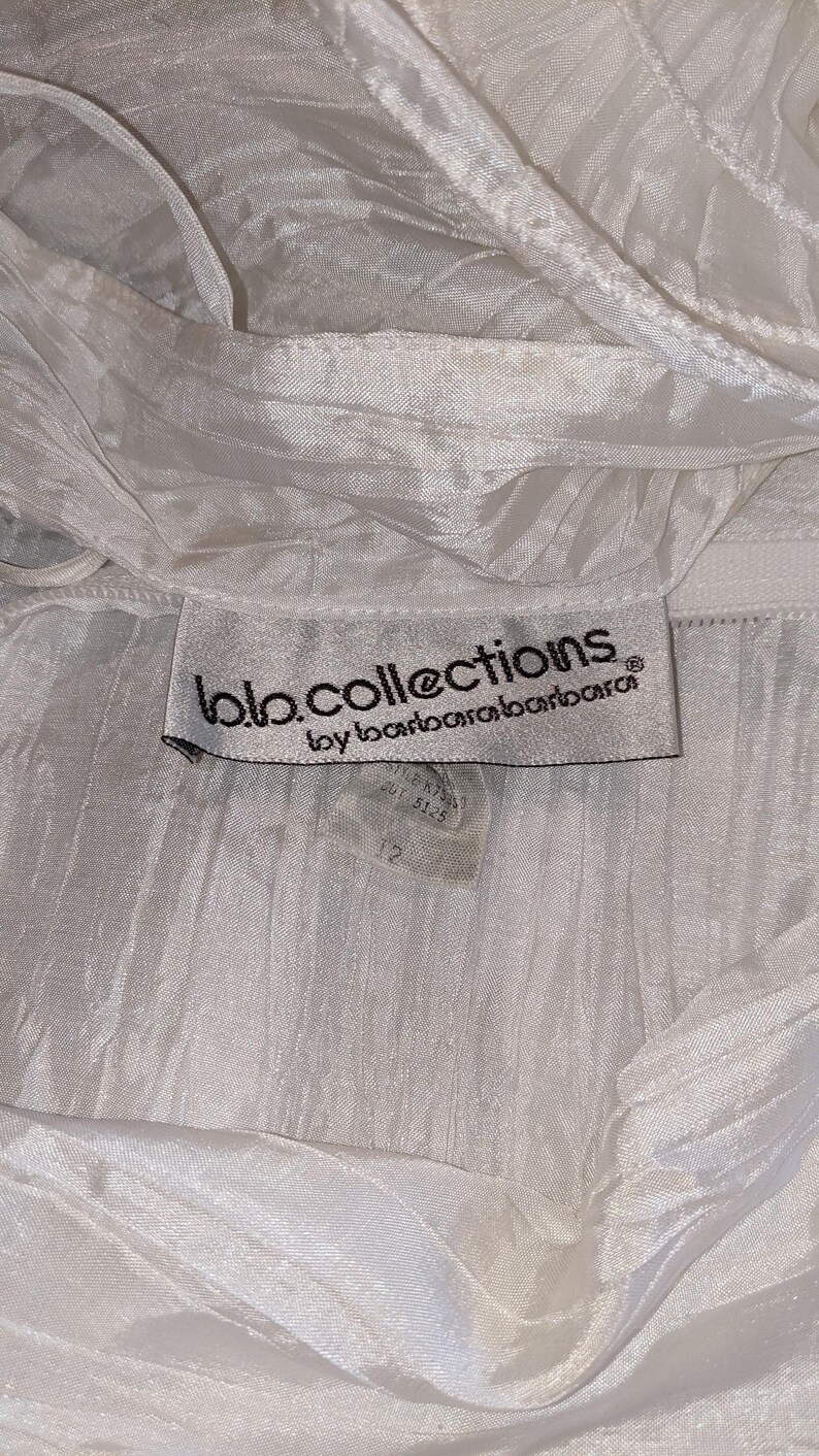 May include: Close-up of a white garment with a textured, pleated fabric. The label reads "bb.collections by barbarabarbarara" with a size 12 tag. The fabric appears to be lightweight and flowing, with a subtle sheen.