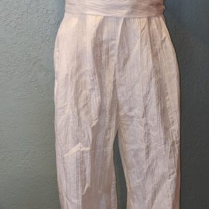 May include: A white jumpsuit with a textured, crinkled fabric. The top features a wrap-style design with ruffled sleeves. The waist is cinched with a matching fabric belt, and the pants have a straight leg. The outfit is displayed on a mannequin.