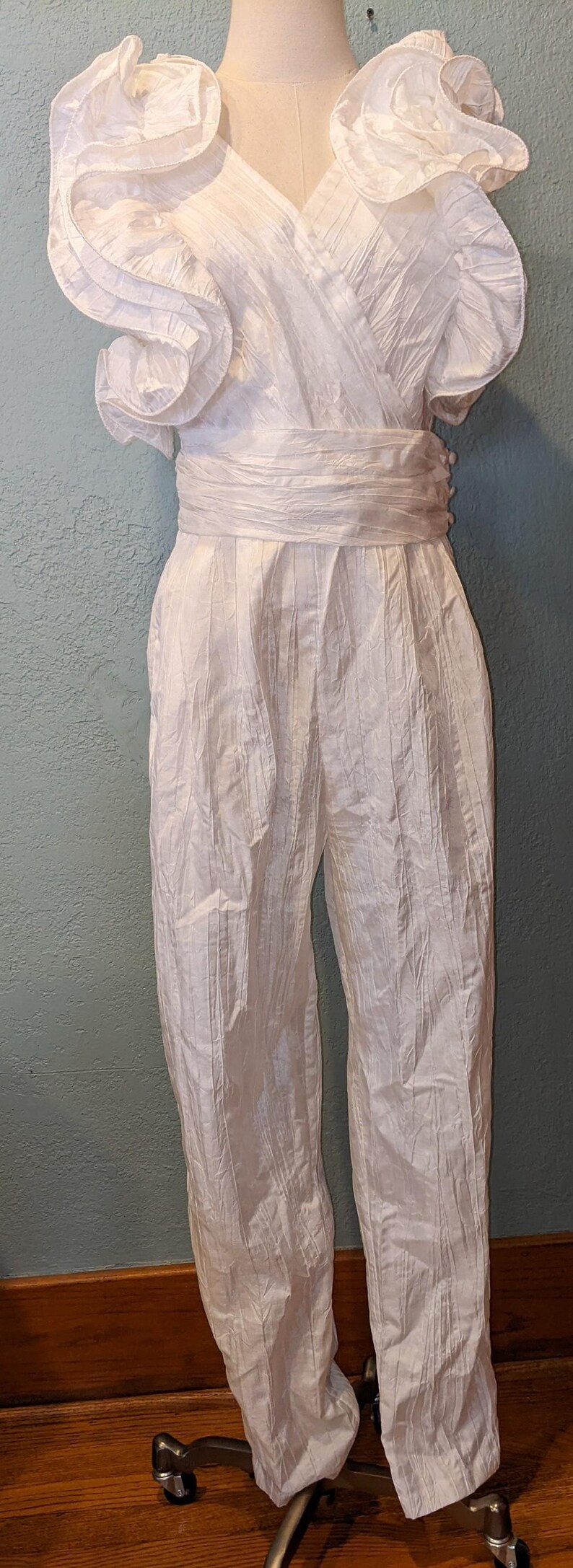 May include: A white jumpsuit with a textured, crinkled fabric. The top features a wrap-style design with ruffled sleeves. The waist is cinched with a matching fabric belt, and the pants have a straight leg. The outfit is displayed on a mannequin.