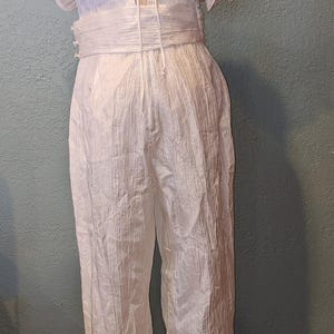 May include: A white jumpsuit with a textured fabric. The top features ruffled sleeves and a low back with tie detailing. The waist is cinched with a matching fabric belt, and the pants are straight-legged.