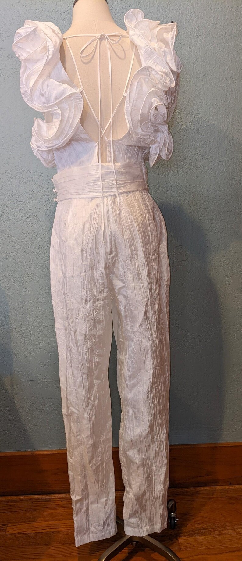 May include: A white jumpsuit with a textured fabric. The top features ruffled sleeves and a low back with tie detailing. The waist is cinched with a matching fabric belt, and the pants are straight-legged.