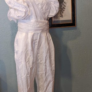 May include: A vintage white jumpsuit with a V-neck, ruffled shoulders, and a wide waist sash. The jumpsuit is displayed on a mannequin. A framed black and white print hangs in the background.