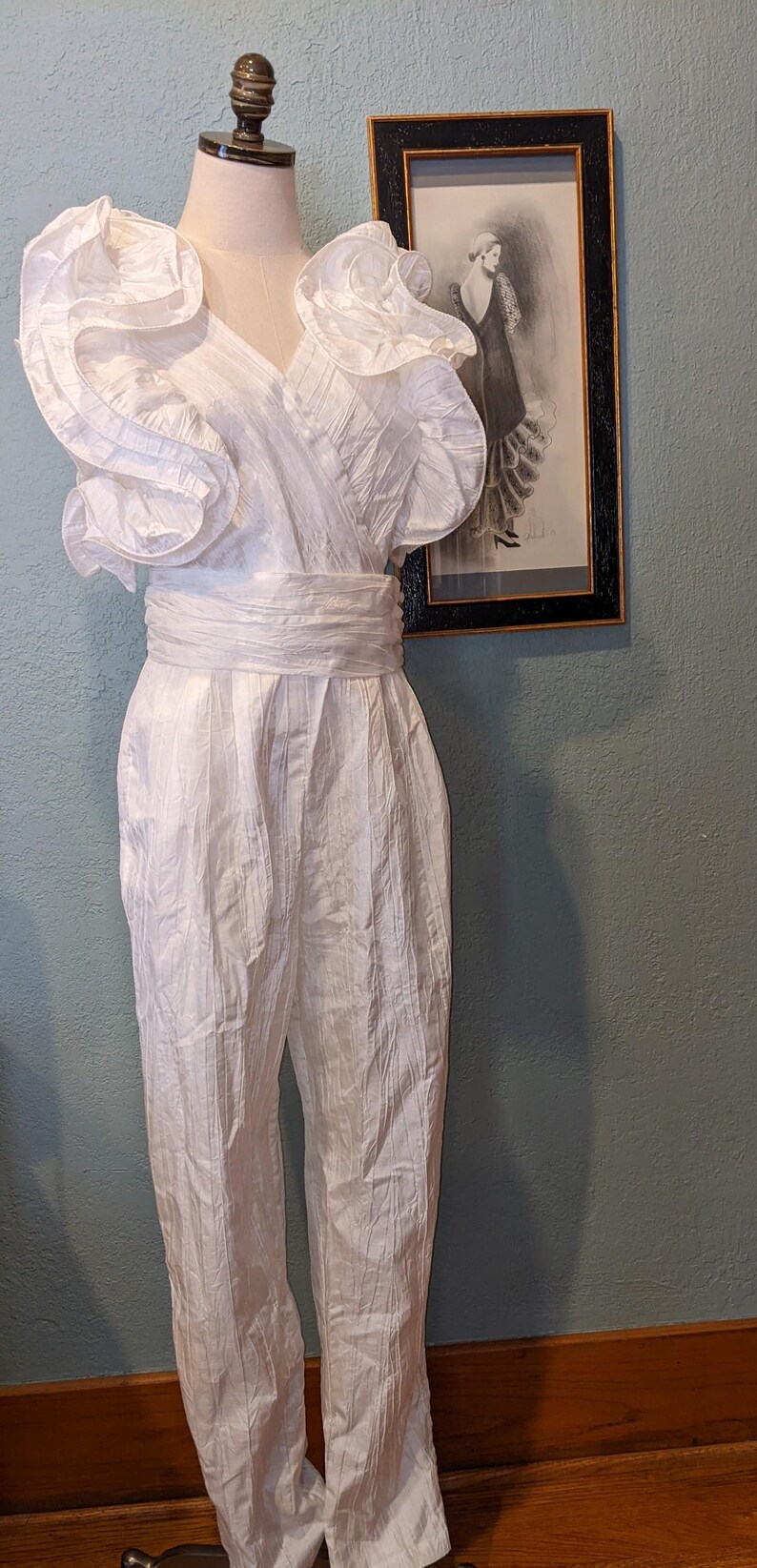 May include: A vintage white jumpsuit with a V-neck, ruffled shoulders, and a wide waist sash. The jumpsuit is displayed on a mannequin. A framed black and white print hangs in the background.