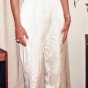 May include: A white jumpsuit with a V-neckline and ruffled sleeves. The jumpsuit has a cinched waist and wide-leg pants. The model is wearing clear heels. The outfit is made of a shiny fabric.