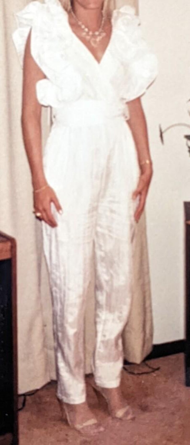 May include: A white jumpsuit with a V-neckline and ruffled sleeves. The jumpsuit has a cinched waist and wide-leg pants. The model is wearing clear heels. The outfit is made of a shiny fabric.