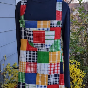 Può includere: Un grembiule patchwork colorato con un'ampia tasca frontale. Il grembiule presenta una varietà di quadrati a quadri e tinta unita in rosso, blu, verde, giallo e bianco. Le cinghie e la tasca sono di colore pieno.