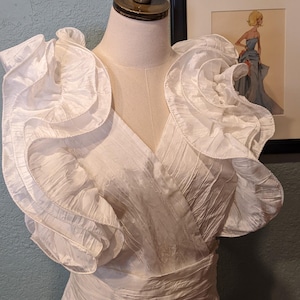May include: A white dress with ruffled sleeves and a V-neck, displayed on a mannequin. The dress has a wrap-style bodice and a defined waist. A framed illustration of a woman in a blue dress is visible in the background.