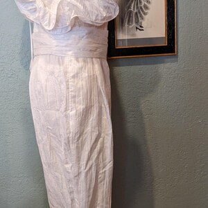 May include: A white, vintage-style dress on a mannequin, featuring a ruffled neckline and a fitted waist. The dress has a textured, vertical striped pattern. A framed sketch of a woman in a dress hangs on the wall behind the mannequin.