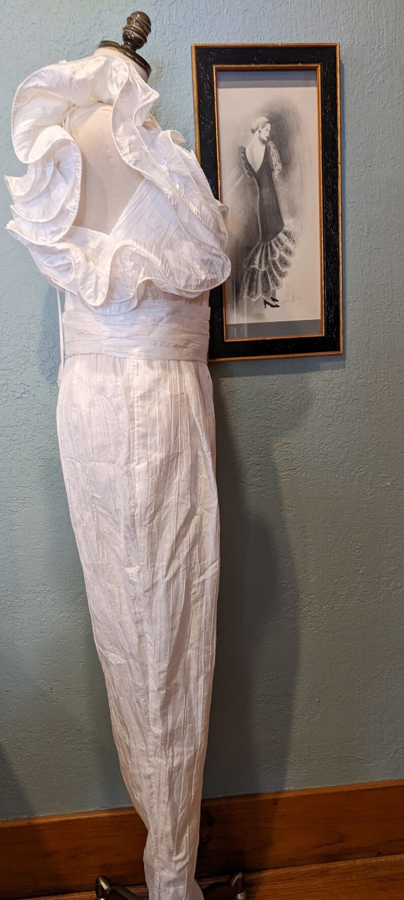 May include: A white, vintage-style dress on a mannequin, featuring a ruffled neckline and a fitted waist. The dress has a textured, vertical striped pattern. A framed sketch of a woman in a dress hangs on the wall behind the mannequin.