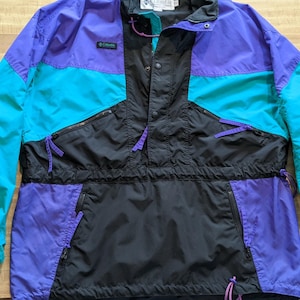 May include: A vintage anorak jacket in a color block design. The jacket features panels of purple, teal, and black. It has a half-zip closure, a front pocket, and the Columbia logo.