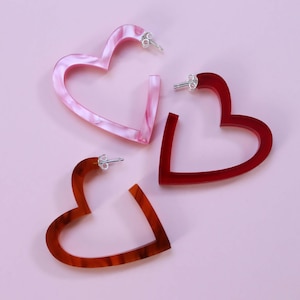 Può includere: Tre orecchini a cerchio a forma di cuore in rosa, rosso e marrone. Gli orecchini sono in acrilico e hanno una finitura lucida.