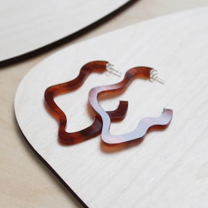 May include: A pair of brown tortoiseshell hoop earrings with a wavy, irregular shape. The earrings are made of a smooth, glossy material and have a silver-toned metal hook fastening.
