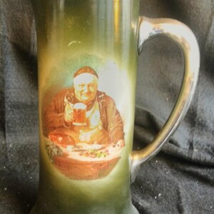 May include: Green ceramic beer stein with a silver handle and rim. The stein features a painted image of a man holding a beer mug. The man is wearing a white hat and apron. The stein has a flared base.