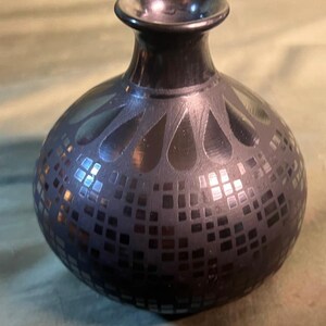 May include: A dark, round vase with a narrow neck and a wide base. The vase is decorated with a pattern of squares and teardrop shapes. The vase is made of a dark material, possibly glass or ceramic.