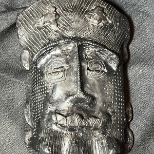 May include: A dark gray, stylized mask with a textured surface. The mask features a prominent nose, a wide, toothy grin, and detailed eyes. The top of the mask has a patterned design, and the overall aesthetic suggests a handcrafted, tribal art piece.