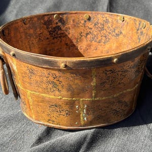 May include: A copper-colored, round container with a dark rim and riveted accents. The container has two metal handles and a textured surface with a grid-like pattern. The overall aesthetic is rustic and vintage.