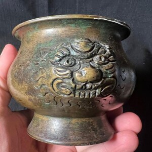 May include: A small, antique bronze bowl with a detailed relief of a mythical creature's face. The bowl has a flared rim and a pedestal base. The bronze has a weathered, aged patina with hints of green oxidation.