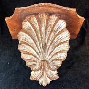 May include: A wooden wall shelf with a seashell design. The shelf is made of wood with a distressed finish. The seashell is painted white and brown, and the shelf is approximately 12 inches wide.