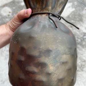 May include: A vintage-style metal vase shaped like a drawstring pouch. The vase has a tarnished, aged appearance with a dark brown and bronze color palette. A twisted rope detail secures the top, adding to its rustic charm.