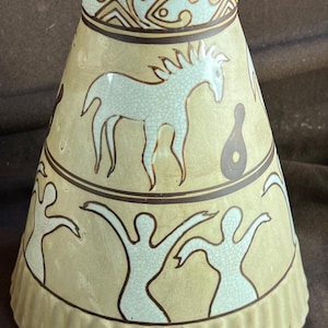 May include: A ceramic vase with a flared shape, featuring a light yellow background and brown accents. The vase is decorated with white horse and human figures, and geometric patterns. The rim is brown.