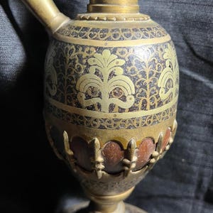 May include: An antique brass hookah with intricate floral and geometric patterns. The hookah features a bulbous body, a curved spout, and a decorative base. The metal has a tarnished, aged appearance, with accents of gold and brown.