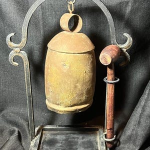 May include: A vintage brass bell with a wooden mallet, suspended from a wrought iron stand. The bell has a cylindrical shape with a rounded top and a loop for hanging. The mallet is brown with a circular head.