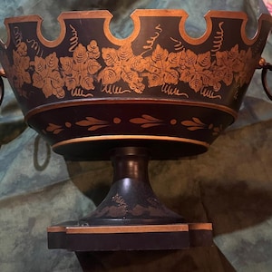 May include: A black decorative bowl with gold accents and a pedestal base. The bowl features a gold grape vine design around the rim and on the sides. Two gold ring handles are attached to the sides of the bowl.