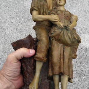 May include: A detailed sculpture of two figures, a man and a woman, in a rustic style. The man wears a hat and holds the woman, both in earth-toned clothing. The base is a dark brown color, and the sculpture has a weathered appearance.