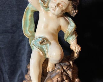 May include: A vintage cherub figurine in cream, gold, and teal. The cherub is posed with one arm raised, draped in a teal cloth, and perched atop a decorative base. The figurine has a matte finish and is likely made of ceramic or plaster.