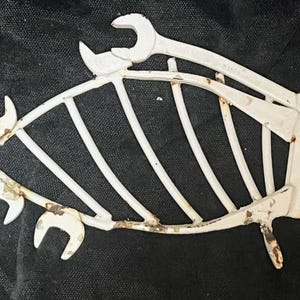 May include: A white metal fish sculpture constructed from wrenches. The fish has a skeletal design with vertical ribs and open jaws. The sculpture has a distressed, rustic appearance, with visible rust.