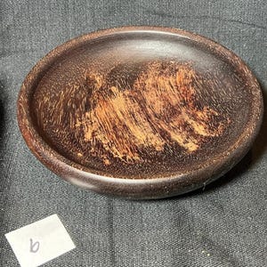 May include: A small, oval-shaped wooden bowl with a dark brown exterior and a lighter, reddish-brown interior. The wood grain is visible, creating a textured appearance. The bowl is likely handmade and suitable for holding small items.