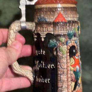 May include: A ceramic beer stein featuring a relief design of a castle tower with a red roof and a figure in an archway. The stein has a metal lid, a textured handle, and black panels with the German text "gute Geister treiber" and "AUCH DIE FR".