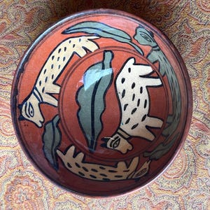 May include: A round, hand-painted ceramic bowl with a reddish-orange background. The bowl features a repeating pattern of stylized, cream-colored animals with black spots and green leaf-like shapes. The rim is a darker shade of brown.