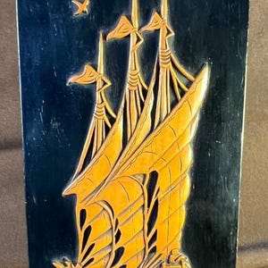 May include: A rectangular, dark blue wooden wall panel with a carved relief of a sailing ship. The ship has three masts with large, golden sails. Two birds are flying above the ship. The artwork is a decorative piece.