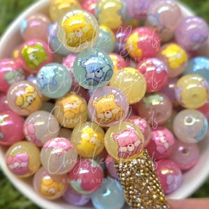 May include: A collection of colorful, translucent round beads with Care Bear designs in a white bowl. The beads are in shades of pink, yellow, blue, and purple. A gold-colored, jeweled pen is in the foreground.