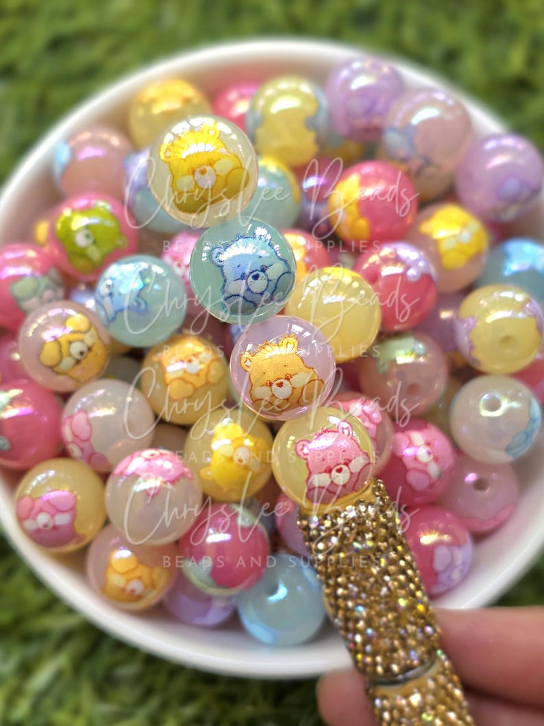 May include: A collection of colorful, translucent round beads with Care Bear designs in a white bowl. The beads are in shades of pink, yellow, blue, and purple. A gold-colored, jeweled pen is in the foreground.