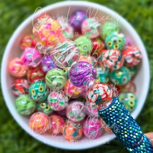 May include: A white bowl filled with colorful, patterned beads. The beads are various colors, including pink, green, orange, and purple. A blue pen with rhinestones is holding a bead. The text "Chryslee Beads" is visible.