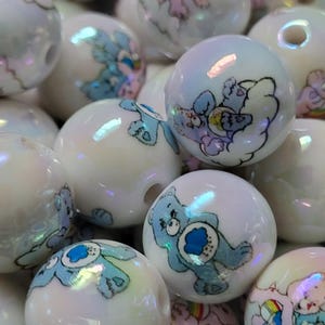 May include: A collection of iridescent white round beads. Each bead features a cartoon bear design in shades of blue, pink, and yellow. The beads have a glossy finish and are suitable for jewelry making or crafting projects.