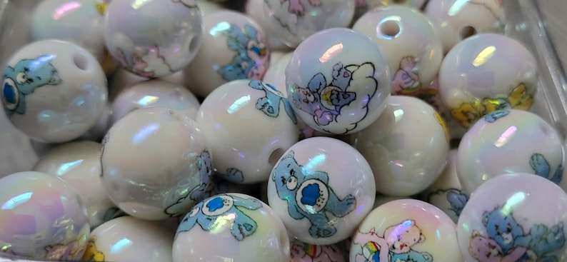 May include: A collection of iridescent white round beads. Each bead features a cartoon bear design in shades of blue, pink, and yellow. The beads have a glossy finish and are suitable for jewelry making or crafting projects.