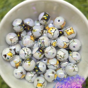 May include: A collection of round, iridescent white beads featuring various cartoon cat illustrations in orange, black, and grey. Each bead has a central hole for stringing. The beads are contained within a white bowl, with the text Chryslee Beads visible.