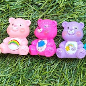 May include: A row of eight colorful bear figurines in various colors, including brown, yellow, pink, purple, teal, green, and blue. Each bear has a unique belly symbol, such as a heart, rainbow, or musical notes. The bears are arranged on a bed of green grass.