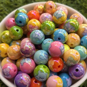 May include: A bowl filled with colorful, round beads. The beads are various colors, including pink, blue, green, and yellow, and feature a cartoon bear design. The beads have a glossy finish and are suitable for jewelry making or crafts.