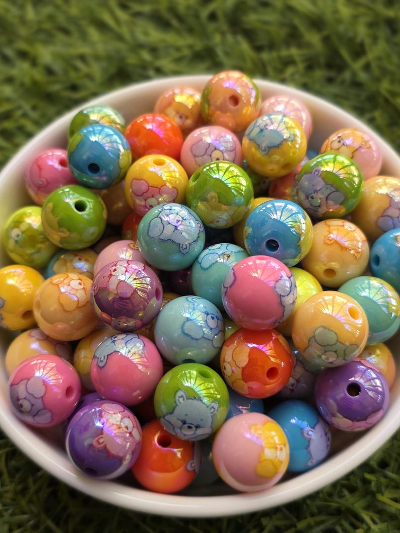 May include: A bowl filled with colorful, round beads. The beads are various colors, including pink, blue, green, and yellow, and feature a cartoon bear design. The beads have a glossy finish and are suitable for jewelry making or crafts.