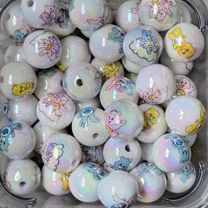 May include: A collection of white, iridescent round beads. Each bead features a colorful Care Bear design. The beads are contained in a clear, square plastic container. The designs include various Care Bears and rainbows.