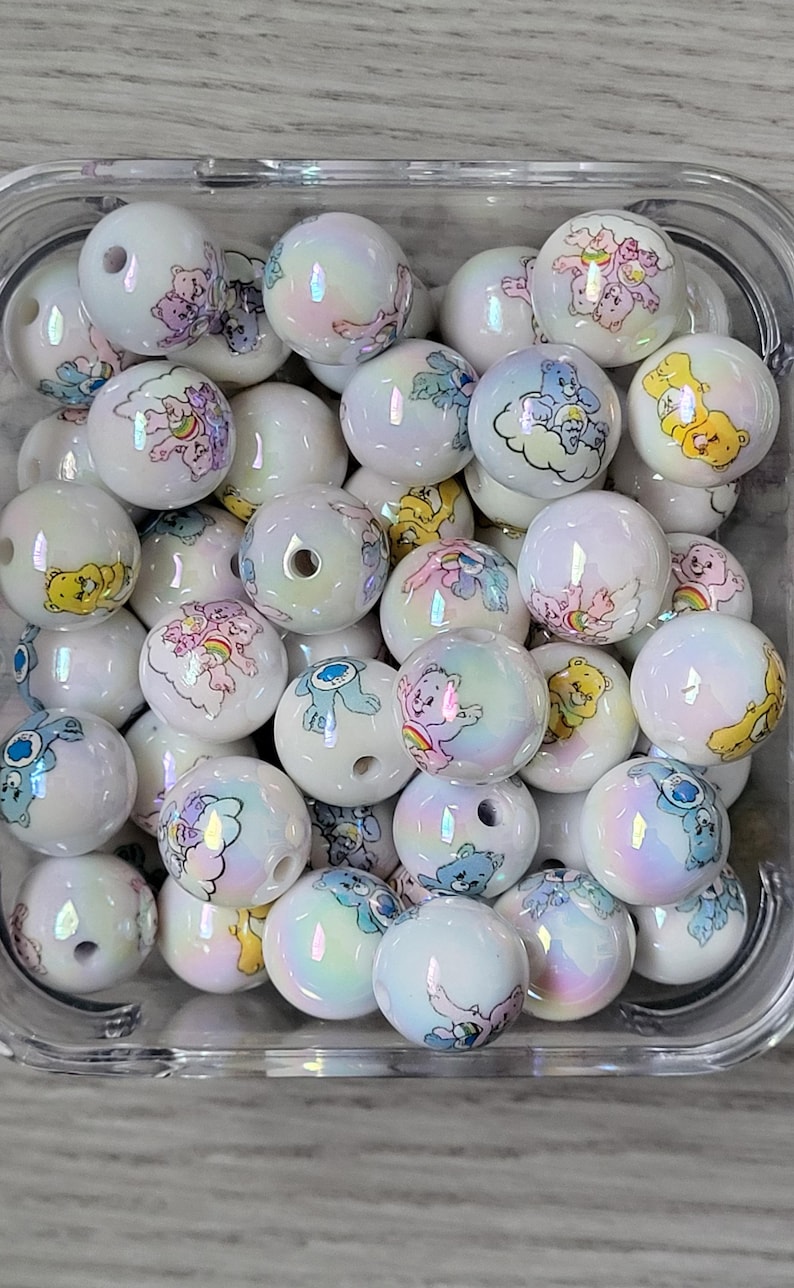May include: A collection of white, iridescent round beads. Each bead features a colorful Care Bear design. The beads are contained in a clear, square plastic container. The designs include various Care Bears and rainbows.